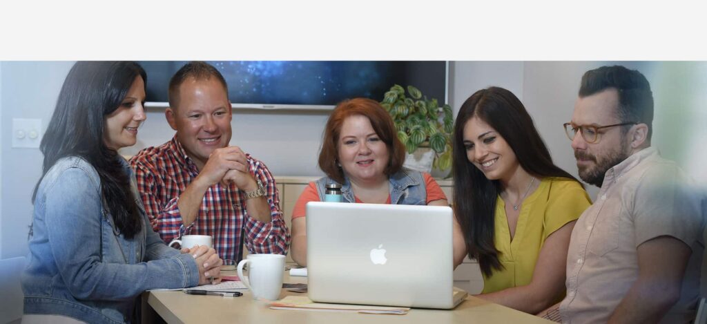 Group of five professionals engaged in a collaborative discussion around a laptop, showcasing teamwork and communication in a modern workspace, relevant to marketing and advertising strategies.