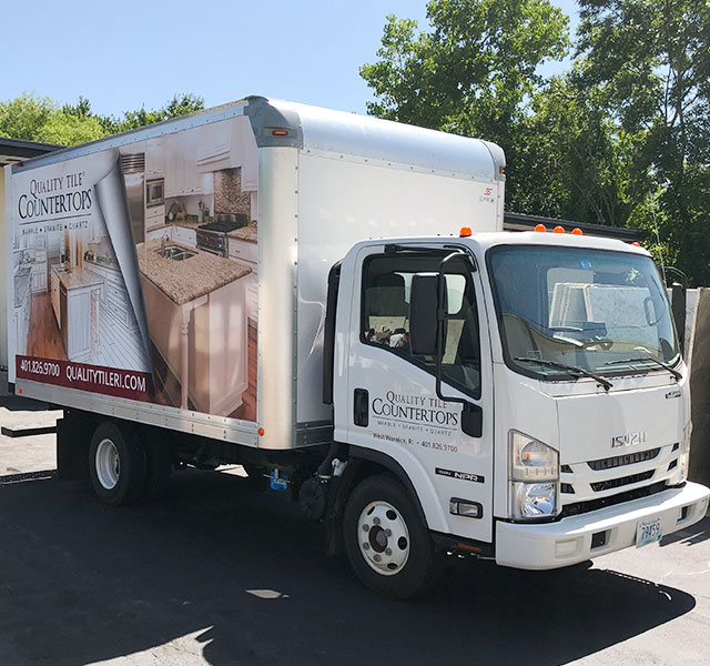 Truck with "Quality Tile Countertops" branding, showcasing kitchen countertops and cabinetry, parked in a green outdoor setting, emphasizing charitable business initiatives.