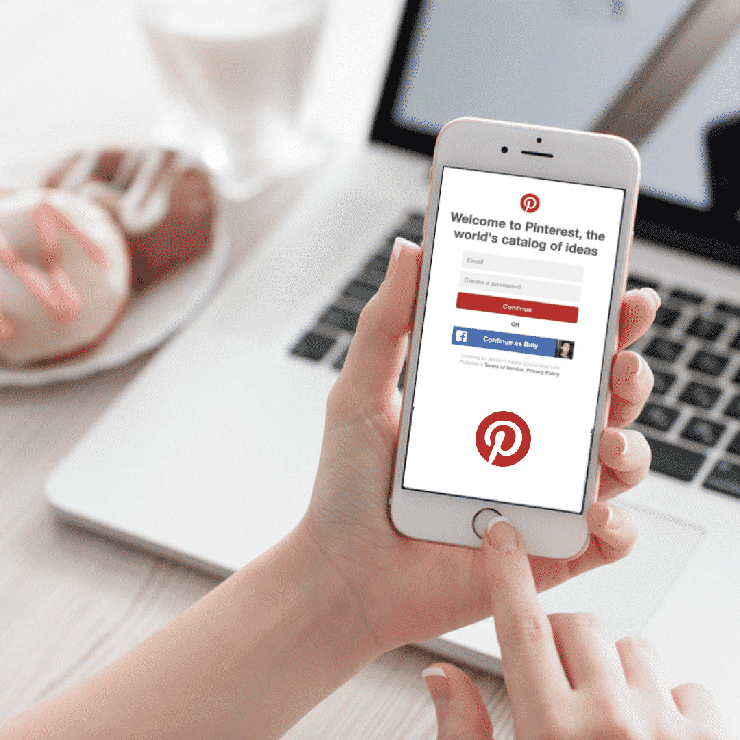 Person holding smartphone displaying Pinterest login screen, with a laptop and dessert in the background, illustrating digital marketing strategies for leveraging Pinterest.