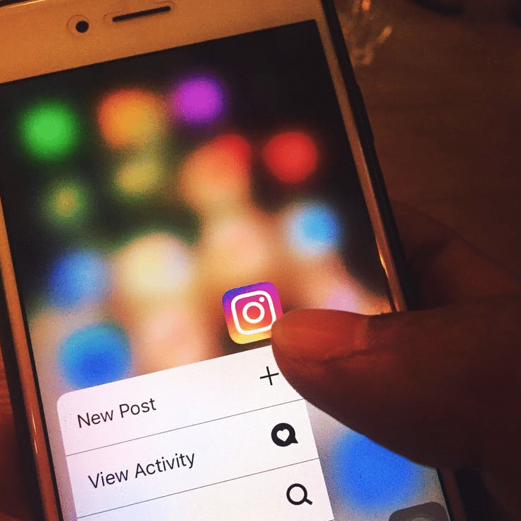 Hand interacting with a smartphone screen displaying the Instagram app icon, featuring options for "New Post" and "View Activity," amidst a blurred colorful background, illustrating social media engagement strategies.