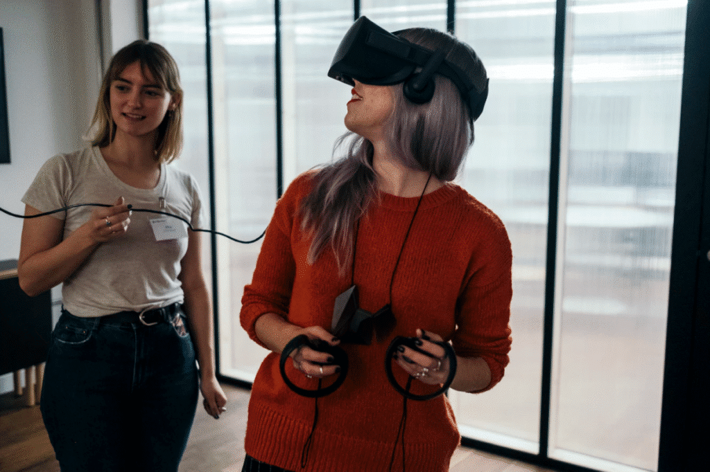 Two women interacting in a modern indoor setting; one wearing a virtual reality headset and holding controllers, while the other offers assistance, highlighting the integration of augmented reality in social media marketing strategies.